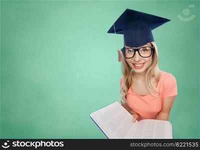 people, education, knowledge and graduation concept - smiling young student woman in mortarboard and eyeglasses with encyclopedia book over green school chalk board background