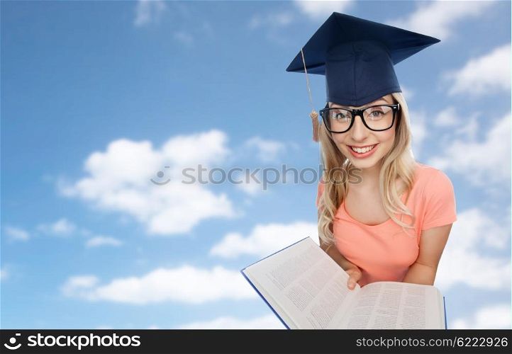 people, education, knowledge and graduation concept - smiling young student woman in mortarboard and eyeglasses with encyclopedia book over blue sky and clouds background