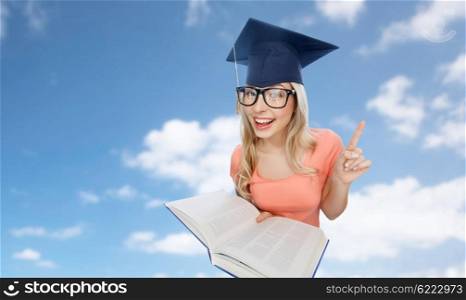 people, education, knowledge and graduation concept - smiling young student woman in mortarboard and eyeglasses with encyclopedia book pointing finger up over blue sky and clouds background