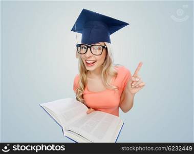 people, education, knowledge and graduation concept - smiling young student woman in mortarboard and eyeglasses with encyclopedia book pointing finger up over gray background