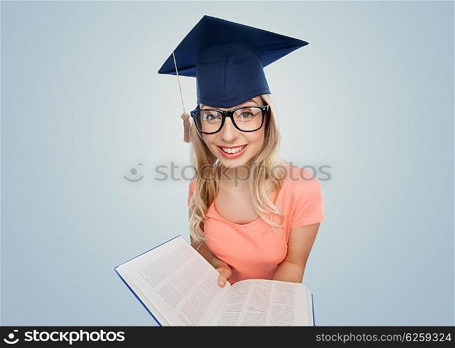 people, education, knowledge and graduation concept - smiling young student woman in mortarboard and eyeglasses with encyclopedia book over gray background