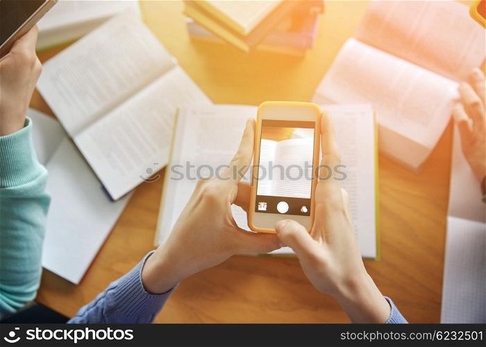 people, education, technology and exam concept - close up of students with smartphones taking picture of books page and making cheat sheet in school library