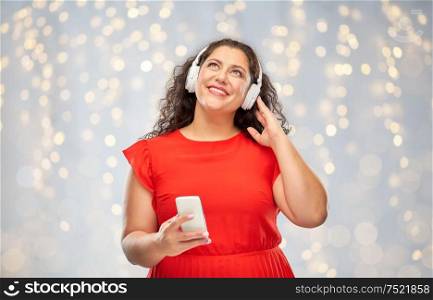 people, electronics and technology concept - happy woman in wireless headphones listening to music on smartphone over festive lights background. woman in headphones listens to music on smartphone