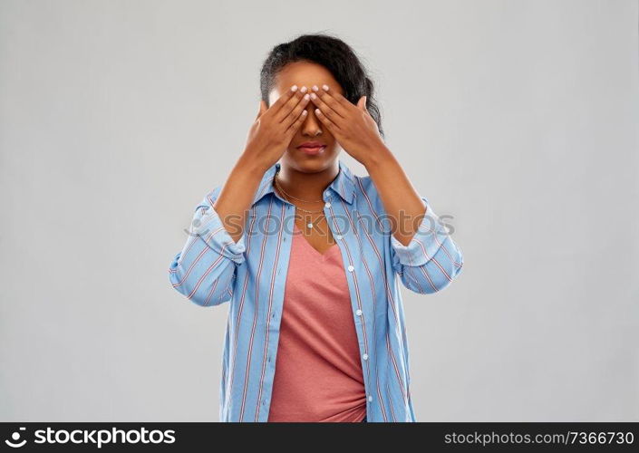 people, emotion and expression concept - african american young woman with eyes closed by hands over grey background. african american woman with eyes closed by hands