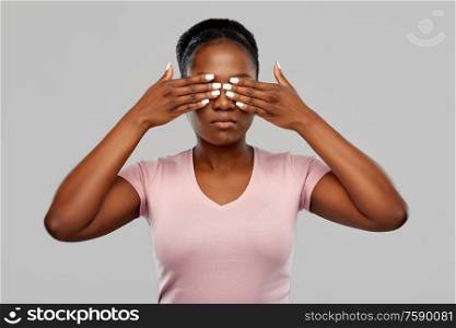 people, emotion and expression concept - african american young woman with eyes closed by hands over grey background. african american woman with eyes closed by hands