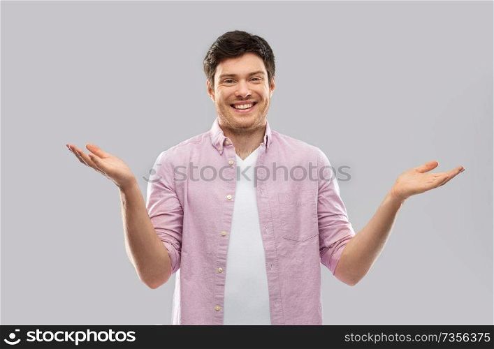 people, emotion and expression concept - happy young man shrugging over grey background. happy young man shrugging over grey background
