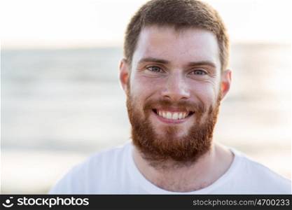 people, emotion and facial expression concept - face of happy smiling young man with red beard on beach