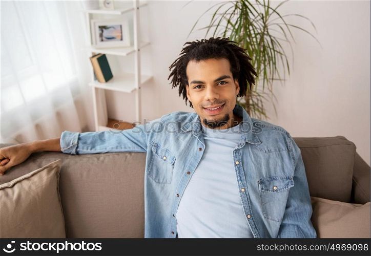 people, emotion and facial expression concept - happy smiling afro american man with dreadlocks sitting on sofa at home. happy smiling afro american man on sofa at home