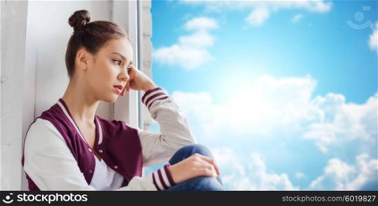 people, emotion and teens concept - sad unhappy pretty teenage girl sitting on windowsill over blue sky and clouds background