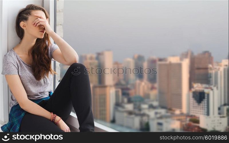people, emotion and teens concept - sad unhappy pretty teenage girl sitting on windowsill over city background