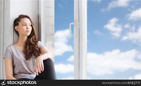 people, emotion and teens concept - sad unhappy pretty teenage girl sitting on windowsill and looking through window over blue sky and clouds background