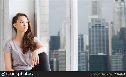 people, emotion and teens concept - sad unhappy pretty teenage girl sitting on windowsill and looking through window over singapore city skyscrapers background