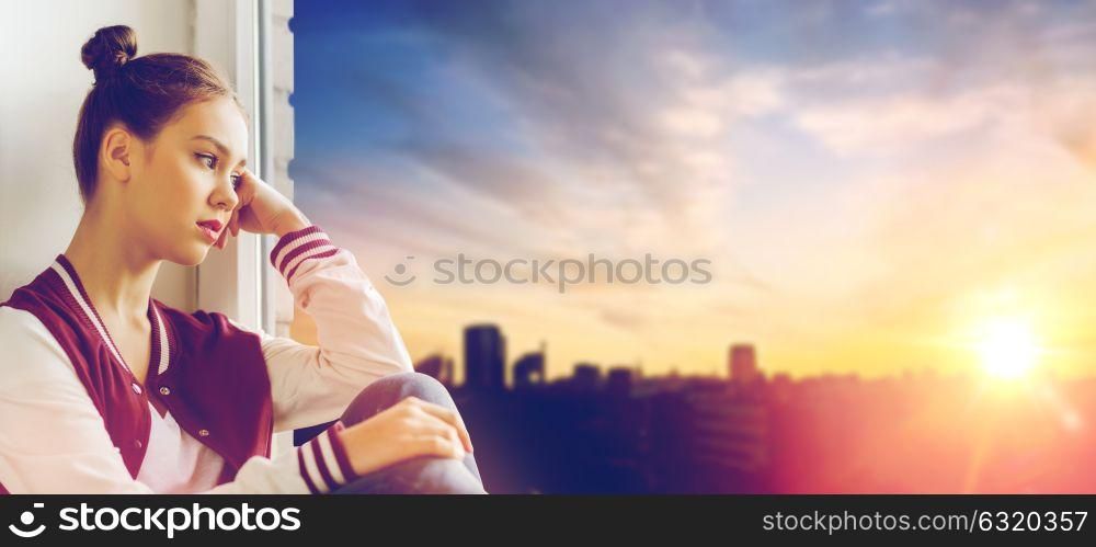 people, emotion and teens concept - sad unhappy pretty teenage girl sitting on windowsill over city background. sad pretty teenage girl sitting on windowsill