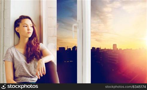 people, emotion and teens concept - sad unhappy pretty teenage girl sitting on windowsill and looking through window over city background. sad pretty teenage girl sitting on windowsill