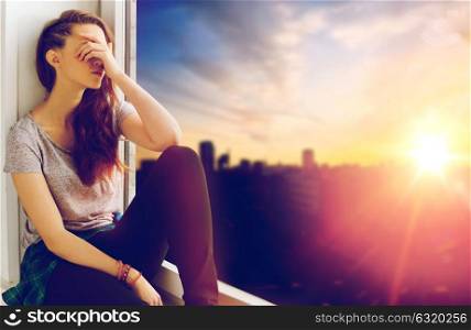 people, emotion and teens concept - sad unhappy teenage girl sitting on windowsill over city background. unhappy teenage girl sitting on windowsill