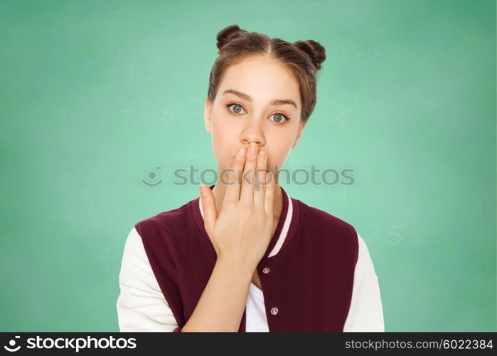 people, emotion, expression, education and teens concept - confused teenage student girl covering her mouth by hand over green school chalk board background