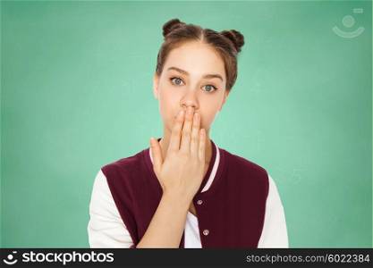 people, emotion, expression, education and teens concept - confused teenage student girl covering her mouth by hand over green school chalk board background