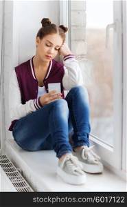 people, emotion, technology and teens concept - sad unhappy pretty teenage girl sitting on windowsill with smartphone and texting