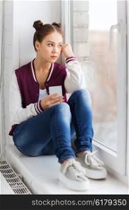 people, emotion, technology and teens concept - sad unhappy pretty teenage girl sitting on windowsill with smartphone and looking to window