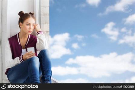 people, emotion, technology and teens concept - sad unhappy pretty teenage girl sitting on windowsill with smartphone and looking through window over blue sky and clouds background