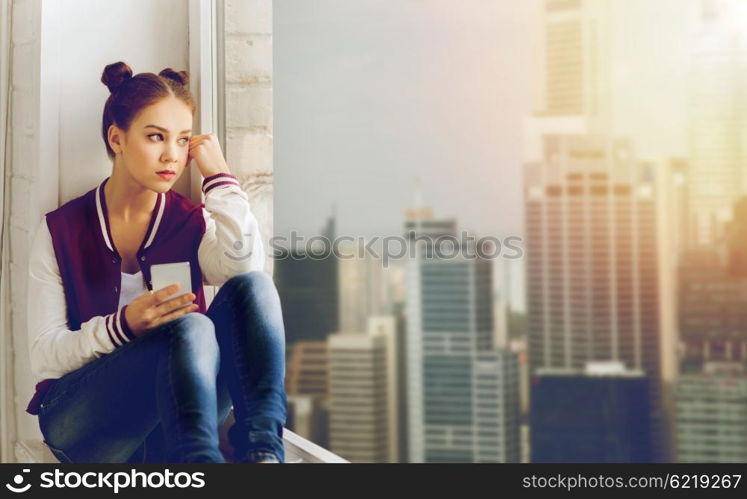 people, emotion, technology and teens concept - sad unhappy pretty teenage girl sitting on windowsill with smartphone and looking to window