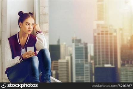 people, emotion, technology and teens concept - sad unhappy pretty teenage girl sitting on windowsill with smartphone and looking to window