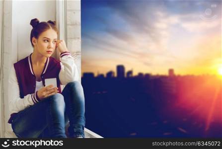 people, emotion, technology and teens concept - sad unhappy pretty teenage girl sitting on windowsill with smartphone and looking through window over city background. teenage girl sitting on windowsill with smartphone