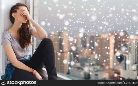 people, emotion, winter, christmas and teens concept - sad unhappy pretty teenage girl sitting at window over city background and snow