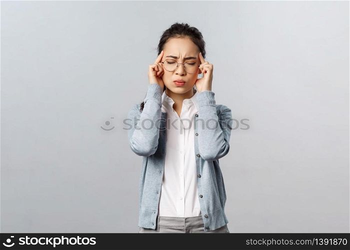People, emotions and lifestyle concept. Troubled young woman in glasses, close eyes and grimacing from pain, touch temples feeling severe headache, suffer painful migraine, grey background.. People, emotions and lifestyle concept. Troubled young woman in glasses, close eyes and grimacing from pain, touch temples feeling severe headache, suffer painful migraine, grey background