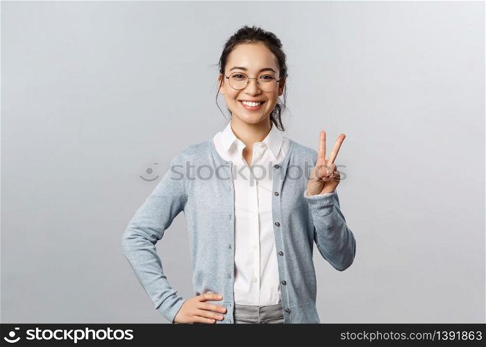 People, emotions and lifestyle concept. Upbeat optimistic asian woman in glasses, school teacher or tutor show peace sign and smiling friendly, express positive cheerful emotion, grey background.. People, emotions and lifestyle concept. Upbeat optimistic asian woman in glasses, school teacher or tutor show peace sign and smiling friendly, express positive cheerful emotion, grey background