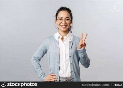 People, emotions and lifestyle concept. Upbeat optimistic asian woman in glasses, school teacher or tutor show peace sign and smiling friendly, express positive cheerful emotion, grey background.. People, emotions and lifestyle concept. Upbeat optimistic asian woman in glasses, school teacher or tutor show peace sign and smiling friendly, express positive cheerful emotion, grey background