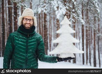 People, emotions, snowy weather and season concept. Happy cheerful young unshaven male dressed in warm clothes, holds fir tree, stands against trees background, enjoys beautiful nature landscapes