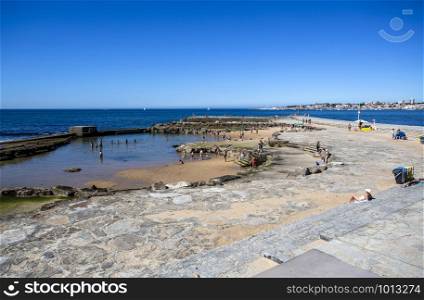 People enjoying the sun and the Atlantic Ocean around a protected pool built on the on the rocks of the Tamariz Beach, in Estoril, Portugal