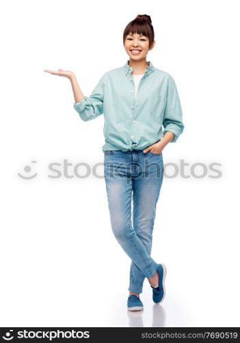 people, ethnicity and advertisement concept - happy asian young woman in cotton shirt and jeans holding something imaginary on her hand over white background. happy asian woman holding something on her hand