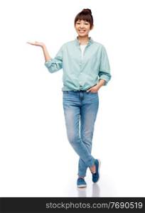people, ethnicity and advertisement concept - happy asian young woman in cotton shirt and jeans holding something imaginary on her hand over white background. happy asian woman holding something on her hand