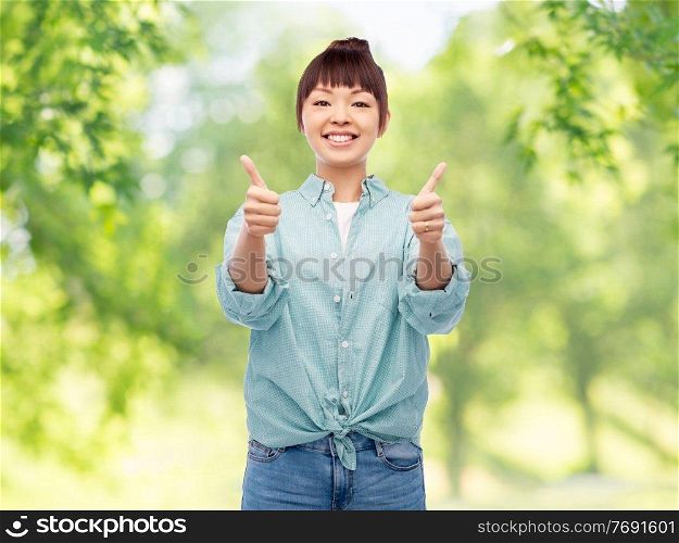people, ethnicity and gesture concept - happy asian young woman in cotton shirt showing thumbs up over green natural background. happy asian woman showing thumbs up over white