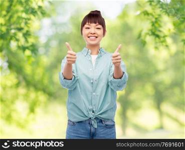 people, ethnicity and gesture concept - happy asian young woman in cotton shirt showing thumbs up over green natural background. happy asian woman showing thumbs up over white