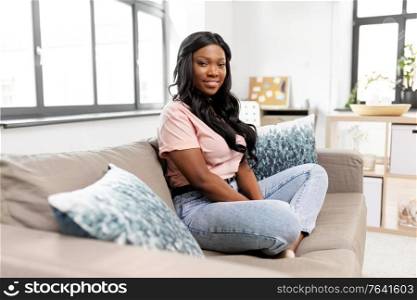 people, ethnicity and portrait concept - happy african american young woman sitting on sofa at home. happy african american young woman at home