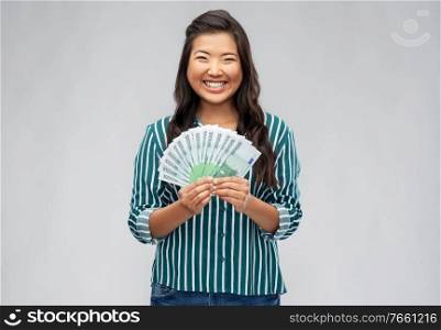 people, ethnicity and portrait concept - happy asian young woman holding hundreds of euro money banknotes over grey background. asian woman with hundred euro money banknotes