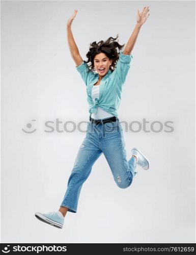 people, ethnicity and portrait concept - happy young woman in turquoise shirt and jeans jumping with hands up over grey background. happy young woman jumping over grey background
