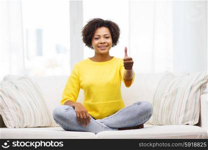 people, ethnicity, gesture and leisure concept - happy african american young woman showing thumbs up and sitting on sofa at home