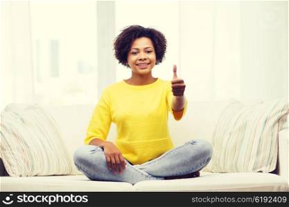 people, ethnicity, gesture and leisure concept - happy african american young woman showing thumbs up and sitting on sofa at home