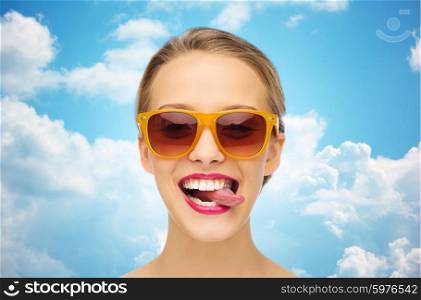 people, expression, joy and fashion concept - smiling young woman in sunglasses with pink lipstick on lips showing tongue over blue sky and clouds background