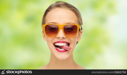 people, expression, joy and fashion concept - smiling young woman in sunglasses with pink lipstick on lips showing tongue over green summer background