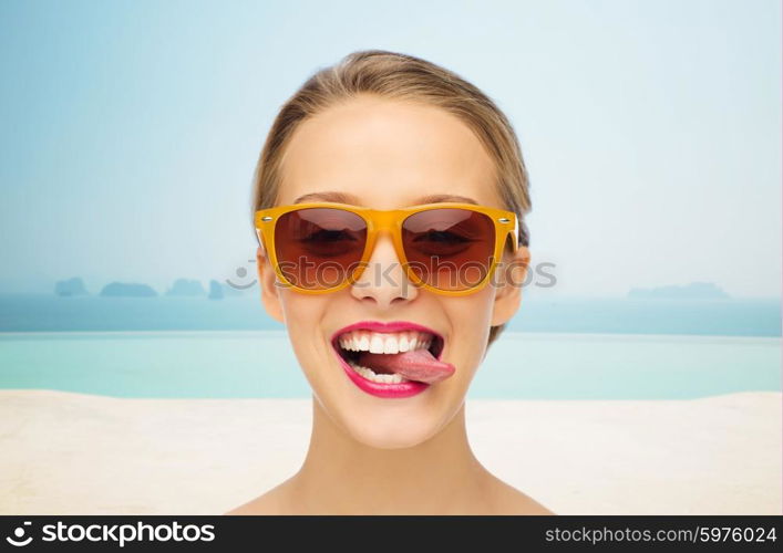 people, expression, vacation, travel and fashion concept - smiling young woman in sunglasses with pink lipstick on lips showing tongue over infinity edge pool background
