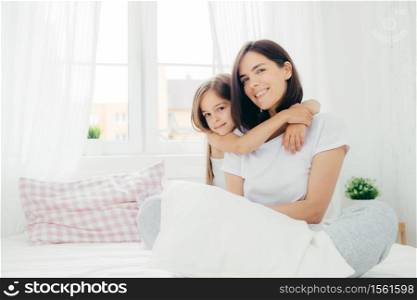 People, family and bedding concept. Cheerful young smiling mother and her daughter embrace each other, have positive expression, wear pyjamas, sit on comfortable bed against window background.