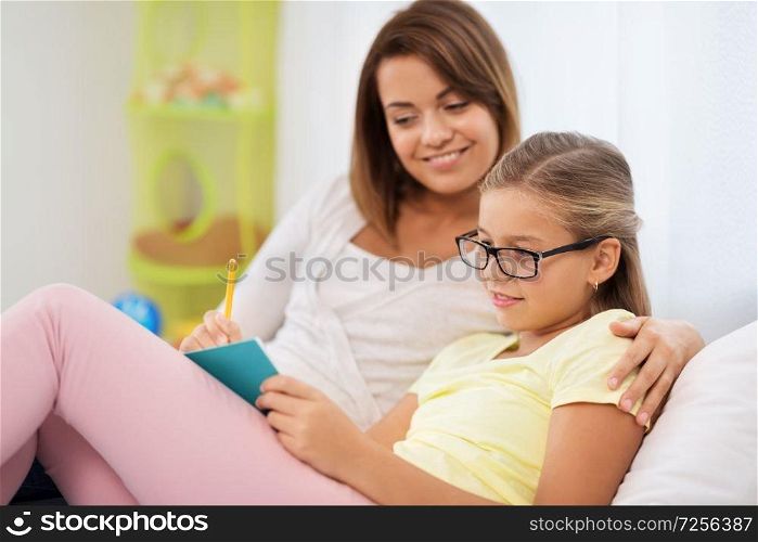 people, family and education concept - happy daughter with mother writing to notebook at home. happy girl with mother writing to notebook at home
