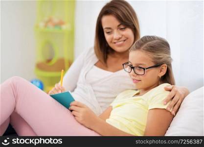 people, family and education concept - happy daughter with mother writing to notebook at home. happy girl with mother writing to notebook at home