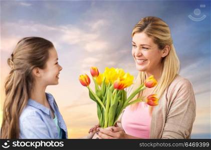people, family and holidays concept - happy daughter giving tulip flowers to her mother over evening sky background. happy daughter giving flowers to mother over sky
