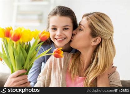 people, family and holidays concept - happy girl giving tulip flowers and kissing her mother at home. happy girl giving flowers to mother at home